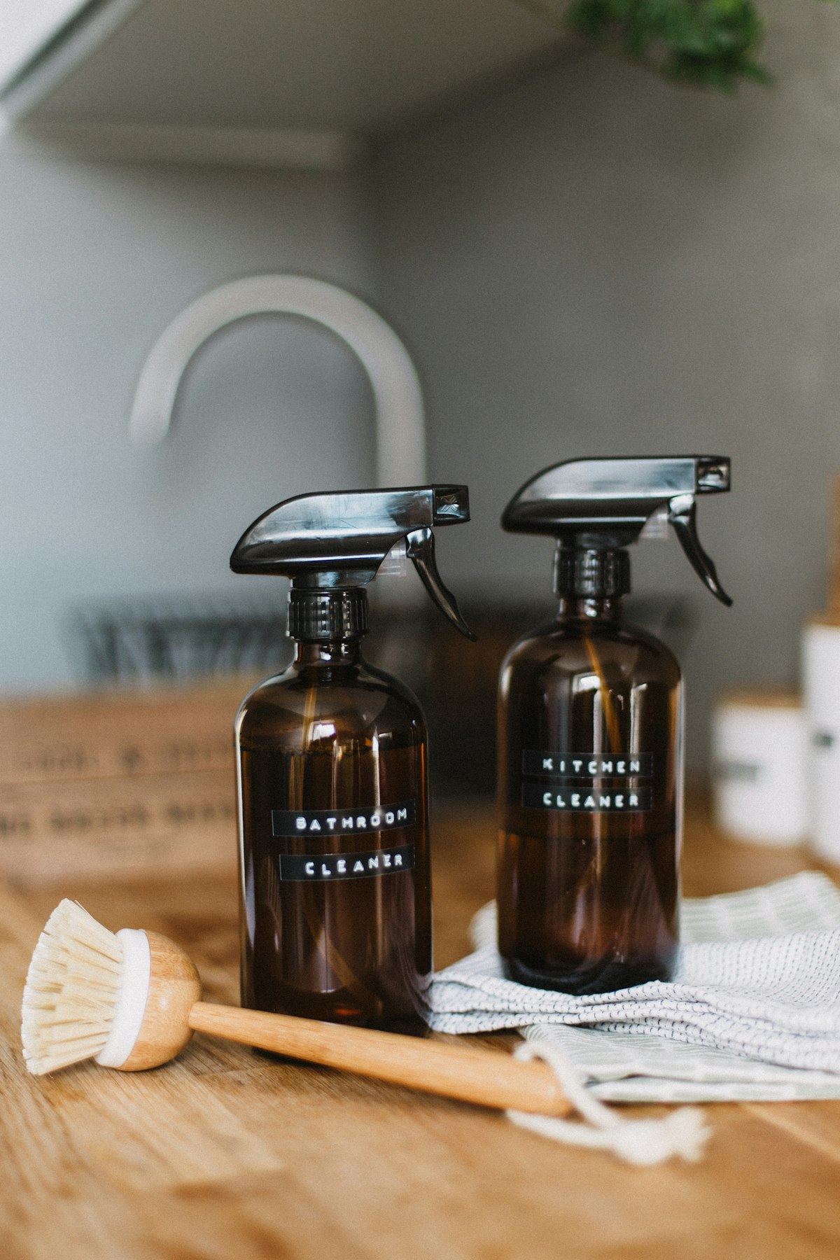 Two brown glass spray bottles and a cleaning brush on a wooden table