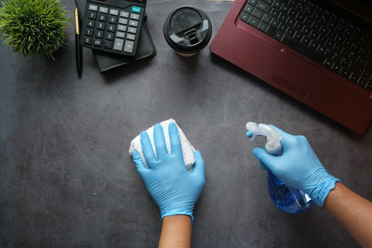 Blue-gloved hands wiping a desk with a cloth and cleaning spray