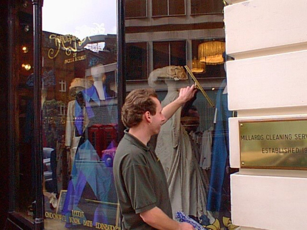 A Millards Cleaning Services window cleaner squeegeeing a shop window in London, beside a brass plaque